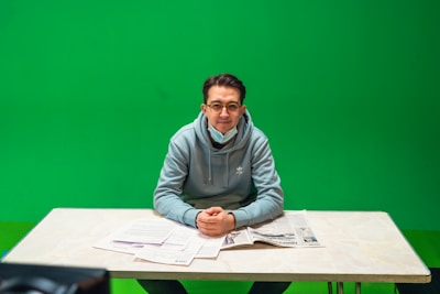 A person wearing glasses and a light blue hoodie is sitting at a table with papers and a newspaper in front. The background is a solid green color, likely used for green screen effects. The person appears to be wearing a face mask pulled down around their neck.