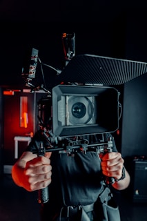 A person holds a large, professional video camera rig with both hands, positioned directly in front of their face. The setting is dimly lit with a soft, red light illuminating parts of the background, suggesting a studio or indoor environment. The camera's lens is prominent, and various technical components and cables are visible.