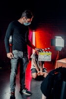 A person wearing a mask is holding a film clapperboard in a dimly lit environment. Another person, sitting on the floor, is bound with hands tied, illuminated by red and white studio lights against a brick wall backdrop.