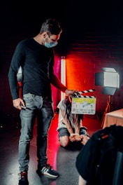 A person wearing a mask is holding a film clapperboard in a dimly lit environment. Another person, sitting on the floor, is bound with hands tied, illuminated by red and white studio lights against a brick wall backdrop.