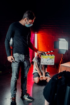 A person wearing a mask is holding a film clapperboard in a dimly lit environment. Another person, sitting on the floor, is bound with hands tied, illuminated by red and white studio lights against a brick wall backdrop.