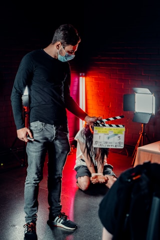 A person wearing a mask is holding a film clapperboard in a dimly lit environment. Another person, sitting on the floor, is bound with hands tied, illuminated by red and white studio lights against a brick wall backdrop.