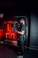 A person is wearing a black t-shirt, gloves, and a harness, operating a professional camera mounted on a stabilizer rig. The background features a dark indoor setting with red lighting, creating a contrasting atmosphere with the person's attire. Equipment and cables are visible on the floor, suggesting a film or video production setting.
