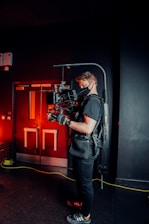 A person is wearing a black t-shirt, gloves, and a harness, operating a professional camera mounted on a stabilizer rig. The background features a dark indoor setting with red lighting, creating a contrasting atmosphere with the person's attire. Equipment and cables are visible on the floor, suggesting a film or video production setting.