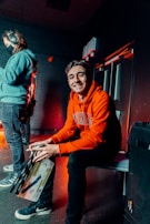 A person wearing an orange hoodie and black pants is sitting on a bench, smiling and holding a clapperboard. Another person, in a teal hoodie and jeans with headphones on, stands to the left, facing away. The room is dimly lit with a reddish ambient light, and exposed brick walls add to the atmosphere.