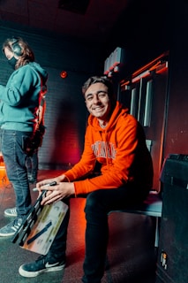 A person wearing an orange hoodie and black pants is sitting on a bench, smiling and holding a clapperboard. Another person, in a teal hoodie and jeans with headphones on, stands to the left, facing away. The room is dimly lit with a reddish ambient light, and exposed brick walls add to the atmosphere.