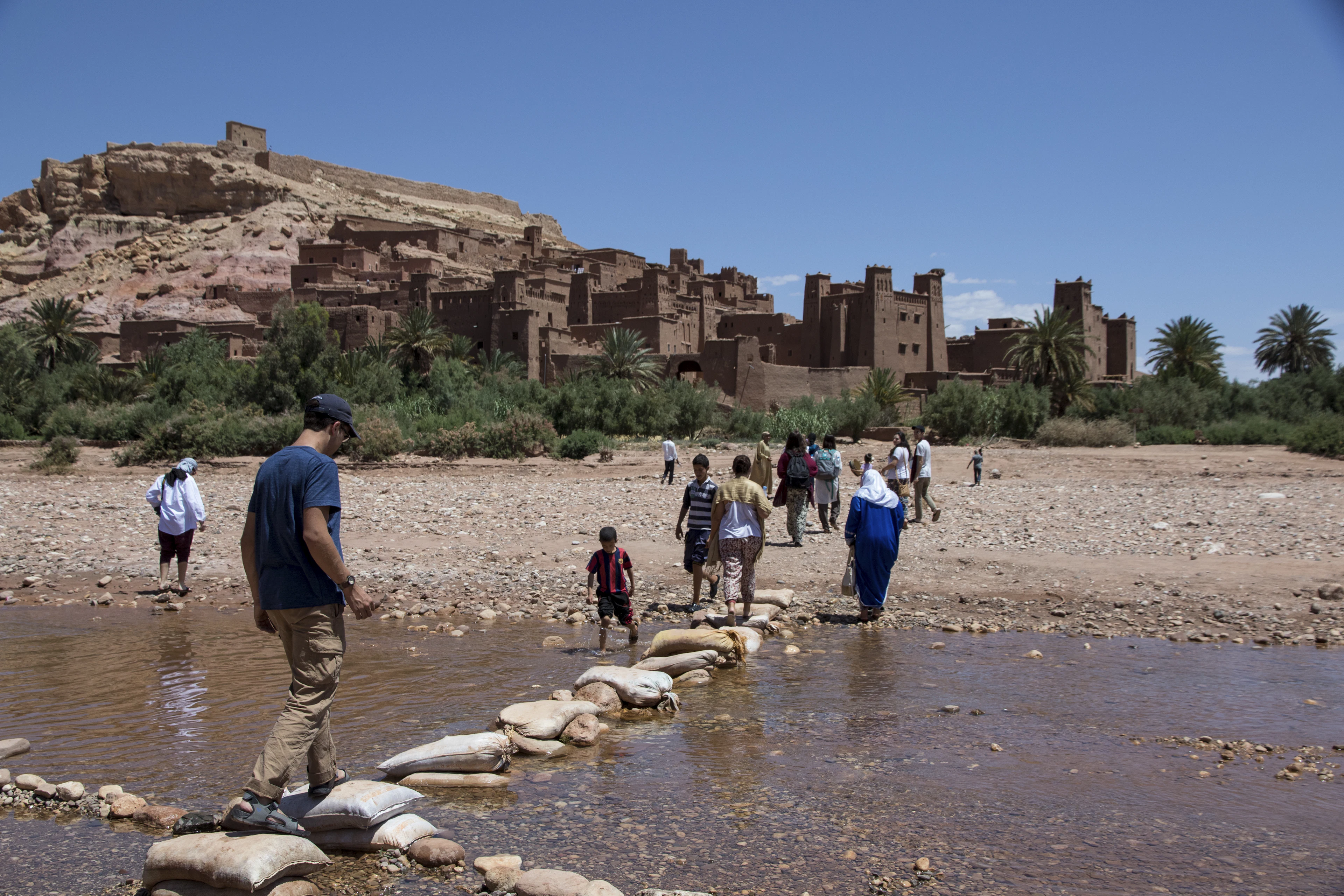 Travellers crossing the river on sandbag stepping stones towards the old earthen ksar on the hill