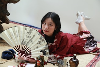 A person in a red kimono is lying on a tatami mat, holding a large decorative fan with images of bamboo and Chinese characters. Surrounding them are various small objects, including a ceramic cat figure, a doll, and several bottles. Their surroundings include traditional Japanese decor.