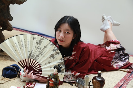 A person in a red kimono is lying on a tatami mat, holding a large decorative fan with images of bamboo and Chinese characters. Surrounding them are various small objects, including a ceramic cat figure, a doll, and several bottles. Their surroundings include traditional Japanese decor.
