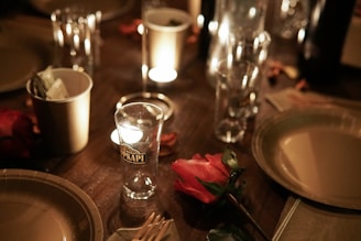 A dimly lit dinner table is set with glassware, including a tall branded glass, surrounded by warm candlelight. There are disposable cups with napkins, a single red rose, plates, and cutlery, creating an intimate dining atmosphere.