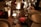 A dimly lit dinner table is set with glassware, including a tall branded glass, surrounded by warm candlelight. There are disposable cups with napkins, a single red rose, plates, and cutlery, creating an intimate dining atmosphere.