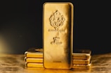 A stack of gold bars is prominently displayed. The forefront features a larger gold bar with intricate engravings, including a lion emblem and inscriptions indicating its weight as one kilo of fine gold. The scene is set against a dark background with a warm, golden glow, highlighting the richness and value of the bars.