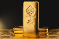 A stack of gold bars is prominently displayed. The forefront features a larger gold bar with intricate engravings, including a lion emblem and inscriptions indicating its weight as one kilo of fine gold. The scene is set against a dark background with a warm, golden glow, highlighting the richness and value of the bars.