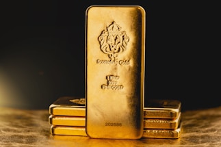 A stack of gold bars is prominently displayed. The forefront features a larger gold bar with intricate engravings, including a lion emblem and inscriptions indicating its weight as one kilo of fine gold. The scene is set against a dark background with a warm, golden glow, highlighting the richness and value of the bars.