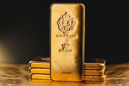 A stack of gold bars is prominently displayed. The forefront features a larger gold bar with intricate engravings, including a lion emblem and inscriptions indicating its weight as one kilo of fine gold. The scene is set against a dark background with a warm, golden glow, highlighting the richness and value of the bars.