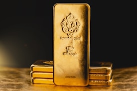 A stack of gold bars is prominently displayed. The forefront features a larger gold bar with intricate engravings, including a lion emblem and inscriptions indicating its weight as one kilo of fine gold. The scene is set against a dark background with a warm, golden glow, highlighting the richness and value of the bars.