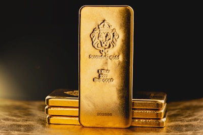 A stack of gold bars is prominently displayed. The forefront features a larger gold bar with intricate engravings, including a lion emblem and inscriptions indicating its weight as one kilo of fine gold. The scene is set against a dark background with a warm, golden glow, highlighting the richness and value of the bars.