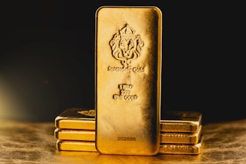 A stack of gold bars is prominently displayed. The forefront features a larger gold bar with intricate engravings, including a lion emblem and inscriptions indicating its weight as one kilo of fine gold. The scene is set against a dark background with a warm, golden glow, highlighting the richness and value of the bars.