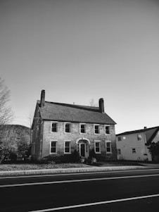 A two-story historic brick house with a classic design, featuring multiple windows and two chimneys. It stands on a neatly maintained lawn across a paved road. Nearby trees and another building are visible under a clear sky.