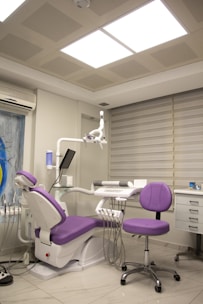 A modern dental clinic room featuring a dentist's chair with purple upholstery and an adjacent matching stool. The room is equipped with dental instruments, an overhead examination light, and a wall-mounted monitor. The walls are adorned with neutral-colored blinds, and there is an air conditioning unit above the equipment. The floor is polished, adding to the clean and sterile atmosphere.