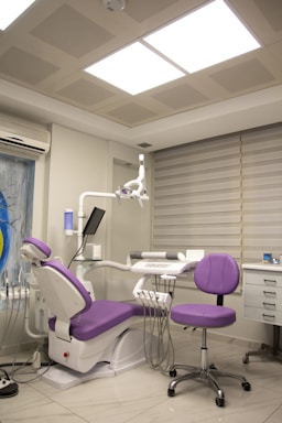 A modern dental clinic room featuring a dentist's chair with purple upholstery and an adjacent matching stool. The room is equipped with dental instruments, an overhead examination light, and a wall-mounted monitor. The walls are adorned with neutral-colored blinds, and there is an air conditioning unit above the equipment. The floor is polished, adding to the clean and sterile atmosphere.