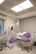 A modern dental clinic room featuring a dentist's chair with purple upholstery and an adjacent matching stool. The room is equipped with dental instruments, an overhead examination light, and a wall-mounted monitor. The walls are adorned with neutral-colored blinds, and there is an air conditioning unit above the equipment. The floor is polished, adding to the clean and sterile atmosphere.