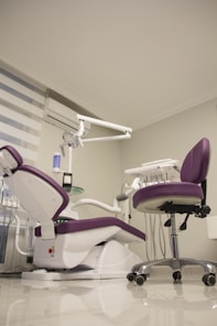 A modern dental office setup features a purple and white dental chair with an attached dental unit, complete with various instruments and a movable overhead lamp. Adjacent, there is a matching purple office chair on wheels. The room has a clean and minimalistic design with white walls and a glossy floor. A window with blinds and an air conditioning unit is visible in the background.