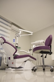 A modern dental office setup features a purple and white dental chair with an attached dental unit, complete with various instruments and a movable overhead lamp. Adjacent, there is a matching purple office chair on wheels. The room has a clean and minimalistic design with white walls and a glossy floor. A window with blinds and an air conditioning unit is visible in the background.