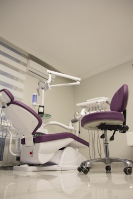 A modern dental office setup features a purple and white dental chair with an attached dental unit, complete with various instruments and a movable overhead lamp. Adjacent, there is a matching purple office chair on wheels. The room has a clean and minimalistic design with white walls and a glossy floor. A window with blinds and an air conditioning unit is visible in the background.