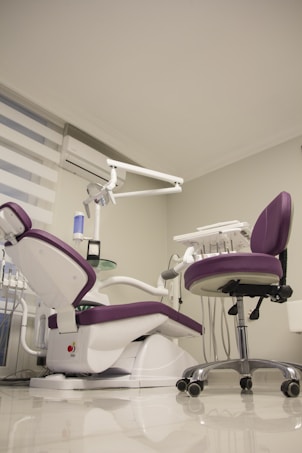 A modern dental office setup features a purple and white dental chair with an attached dental unit, complete with various instruments and a movable overhead lamp. Adjacent, there is a matching purple office chair on wheels. The room has a clean and minimalistic design with white walls and a glossy floor. A window with blinds and an air conditioning unit is visible in the background.