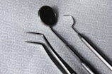 A set of dental instruments is arranged on a fabric with a hexagonal pattern. The tools include a dental mirror, a scaler, forceps, and a pair of tweezers, which are all made of shiny metal.