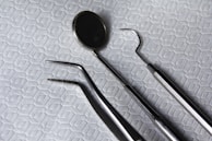 A set of dental instruments is arranged on a fabric with a hexagonal pattern. The tools include a dental mirror, a scaler, forceps, and a pair of tweezers, which are all made of shiny metal.