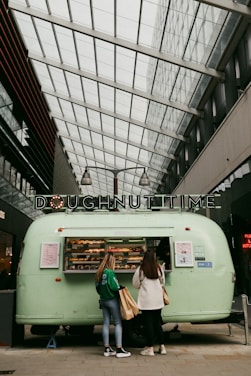 A mint green doughnut food truck is parked indoors under a glass ceiling. Two women are standing in front of it, engaging with the vendor. A variety of colorful doughnuts are displayed in the truck’s window. The sign on top reads 'DOUGHNUT TIME' in bold letters. The setting appears to be a modern urban building with high ceilings and natural light.