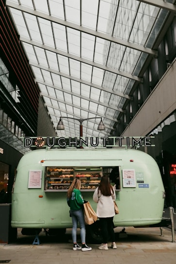 A mint green doughnut food truck is parked indoors under a glass ceiling. Two women are standing in front of it, engaging with the vendor. A variety of colorful doughnuts are displayed in the truck’s window. The sign on top reads 'DOUGHNUT TIME' in bold letters. The setting appears to be a modern urban building with high ceilings and natural light.