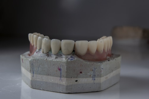 A dental mold featuring a set of artificial teeth is placed on a smooth surface. The mold is crafted from a greyish material with slight discolorations and scribbles. The artificial teeth are aligned neatly, resembling a realistic dental setup with gums depicted in a darker, reddish shade.