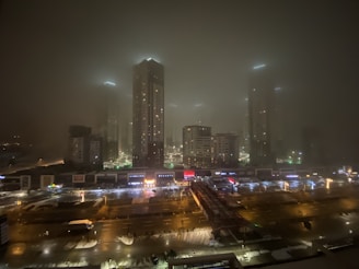 A nighttime urban landscape featuring tall buildings enveloped in fog, lit by artificial lights that create a moody, atmospheric effect. The foreground shows a busy city street with cars and brightly lit shop signs, while the skyscrapers in the background rise into the misty sky.