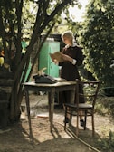 A person with braided hair stands in a garden setting, reading a book near an old typewriter on a rustic table. Sunlight filters through the lush green leaves of the trees above. The scene includes a wooden chair and the ground is covered with pebbles and patches of grass.