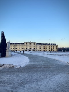 A large historic palace with a symmetrical and ornate facade is set against a clear blue sky. The foreground features a snow-covered path leading towards the building, with a few people walking along it. A statue and hedges are visible on the sides, contributing to the formal layout.