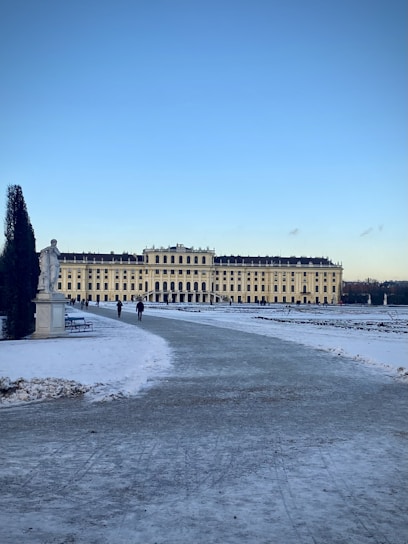 A large historic palace with a symmetrical and ornate facade is set against a clear blue sky. The foreground features a snow-covered path leading towards the building, with a few people walking along it. A statue and hedges are visible on the sides, contributing to the formal layout.