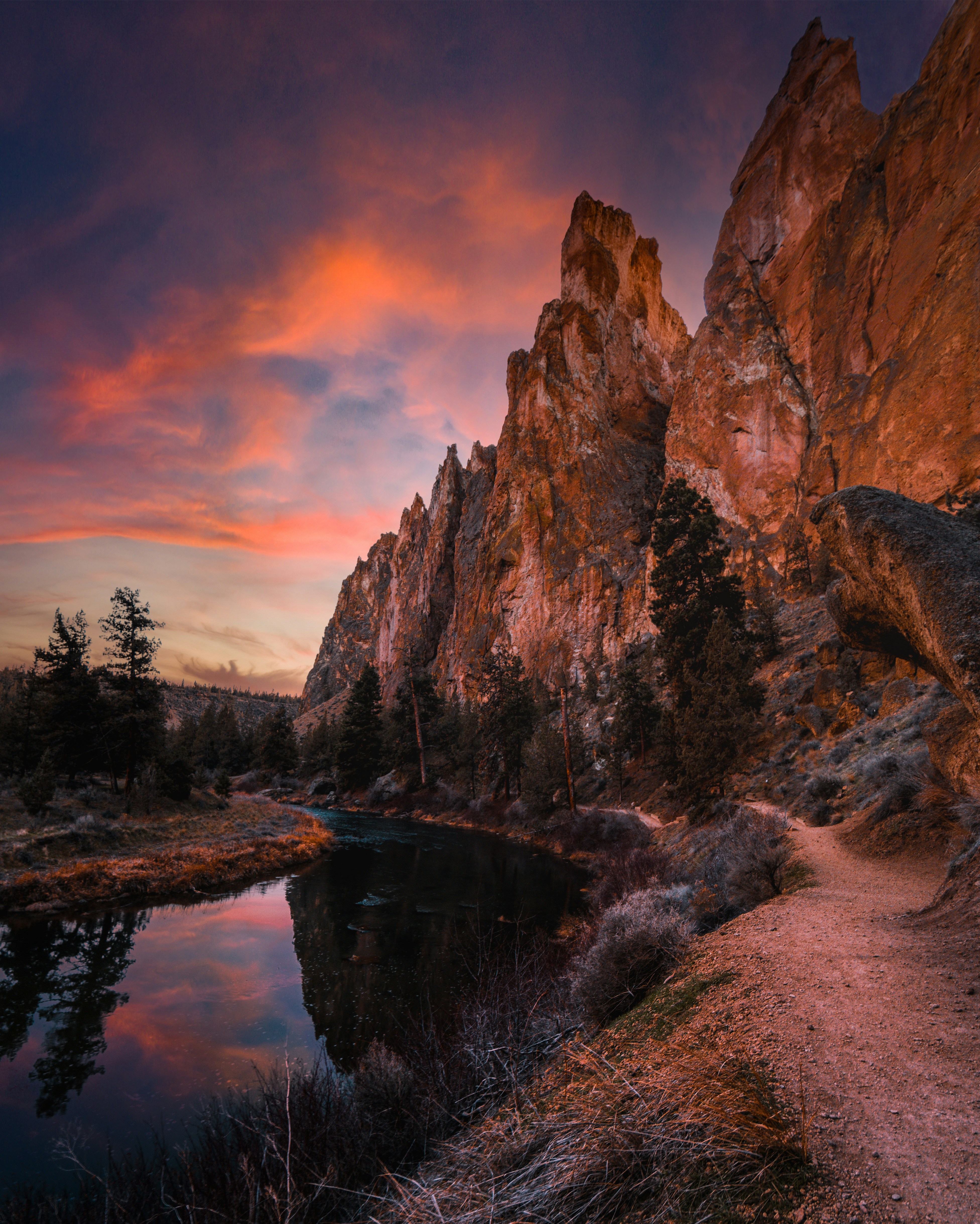 River reflecting a pink sunset below tall rock spires, a dirt trail running along the bank