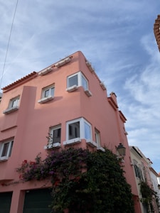 A charming pink building with white window frames and a terracotta-tiled roof sits under a clear blue sky. Lush vines with purple and red flowers climb the facade, adding a quaint and vibrant touch. A traditional street lamp is affixed to the building, enhancing the picturesque atmosphere.