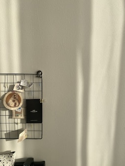 A wall-mounted grid organizer holds various items including a wooden circle with a butterfly design, a black booklet, and several small pieces of paper clipped with colorful paperclips. Soft shadows are cast on the wall, possibly from natural light.