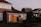 A residential area with a semi-detached house featuring red brick walls and a brown tiled roof. The front door and garage door are painted bright yellow, and the house has two upper-story windows with white lace curtains. The garden is well-maintained with neatly trimmed hedges and lush green grass. In the background, there are additional homes with similar architecture under a clear sky during twilight.