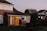 A residential area with a semi-detached house featuring red brick walls and a brown tiled roof. The front door and garage door are painted bright yellow, and the house has two upper-story windows with white lace curtains. The garden is well-maintained with neatly trimmed hedges and lush green grass. In the background, there are additional homes with similar architecture under a clear sky during twilight.