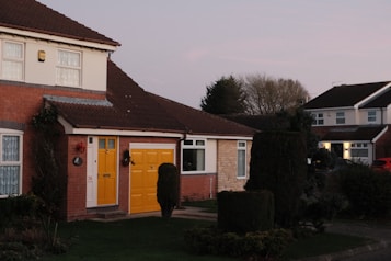 A residential area with a semi-detached house featuring red brick walls and a brown tiled roof. The front door and garage door are painted bright yellow, and the house has two upper-story windows with white lace curtains. The garden is well-maintained with neatly trimmed hedges and lush green grass. In the background, there are additional homes with similar architecture under a clear sky during twilight.