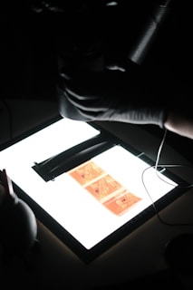 A hand wearing a glove is holding a film negative strip over a lightbox. The film strip contains several images, possibly taken with an analog camera. The surrounding environment is dimly lit, emphasizing the illuminated area of the lightbox.