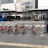 A row of red bicycles lined up at a station on a city street with a backdrop of modern urban buildings. The environment is clean and organized, with a bus stop and a blue bus visible in the background. The foreground features a clear pedestrian pavement with tactile paving.