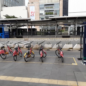 A row of red bicycles lined up at a station on a city street with a backdrop of modern urban buildings. The environment is clean and organized, with a bus stop and a blue bus visible in the background. The foreground features a clear pedestrian pavement with tactile paving.
