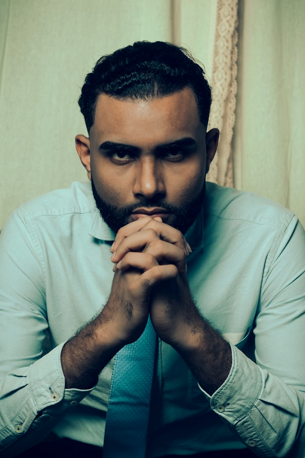 a man in a blue shirt and tie sitting in front of a curtain