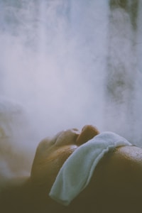 A person is lying down with a white cloth or towel placed over their head, surrounded by mist or steam. The scene suggests a relaxing or therapeutic environment.